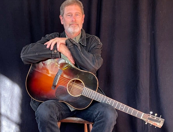 Dana Robinson siting on a stool leaning on his guitar